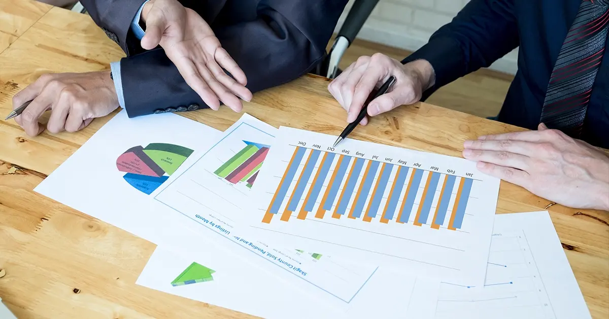 Two professionals in suits analyzing printed bar graphs and pie charts on a wooden desk.