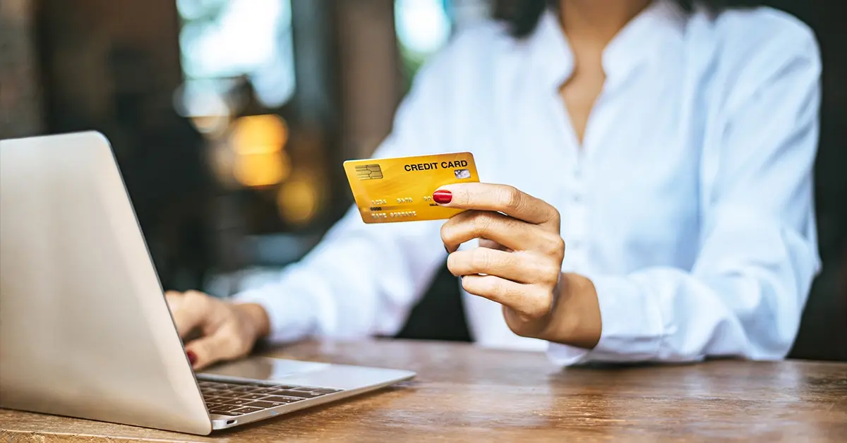 A woman in a white shirt holding a gold credit card while using a laptop on a wooden desk.